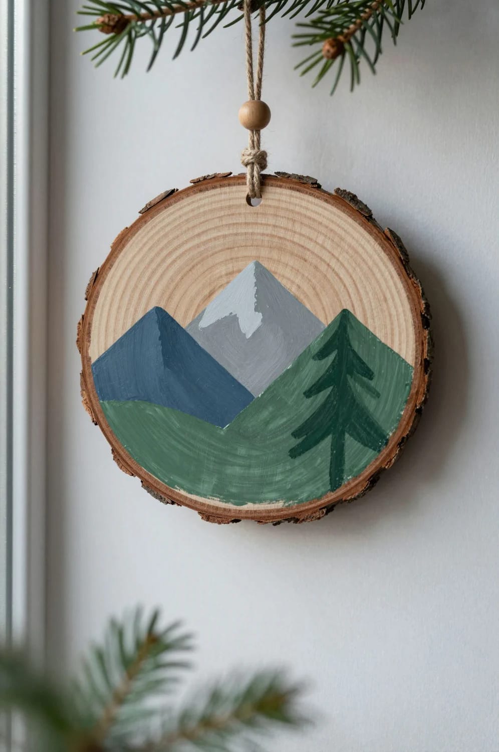 13 Painted Wood Slice Ornaments for a Minimalist Tree 5 A round wood slice ornament featuring a hand-painted minimalist mountain range and pine tree, hanging from a string on a Christmas tree branch, illustrating a simple design idea for painted wood slice ornaments.
