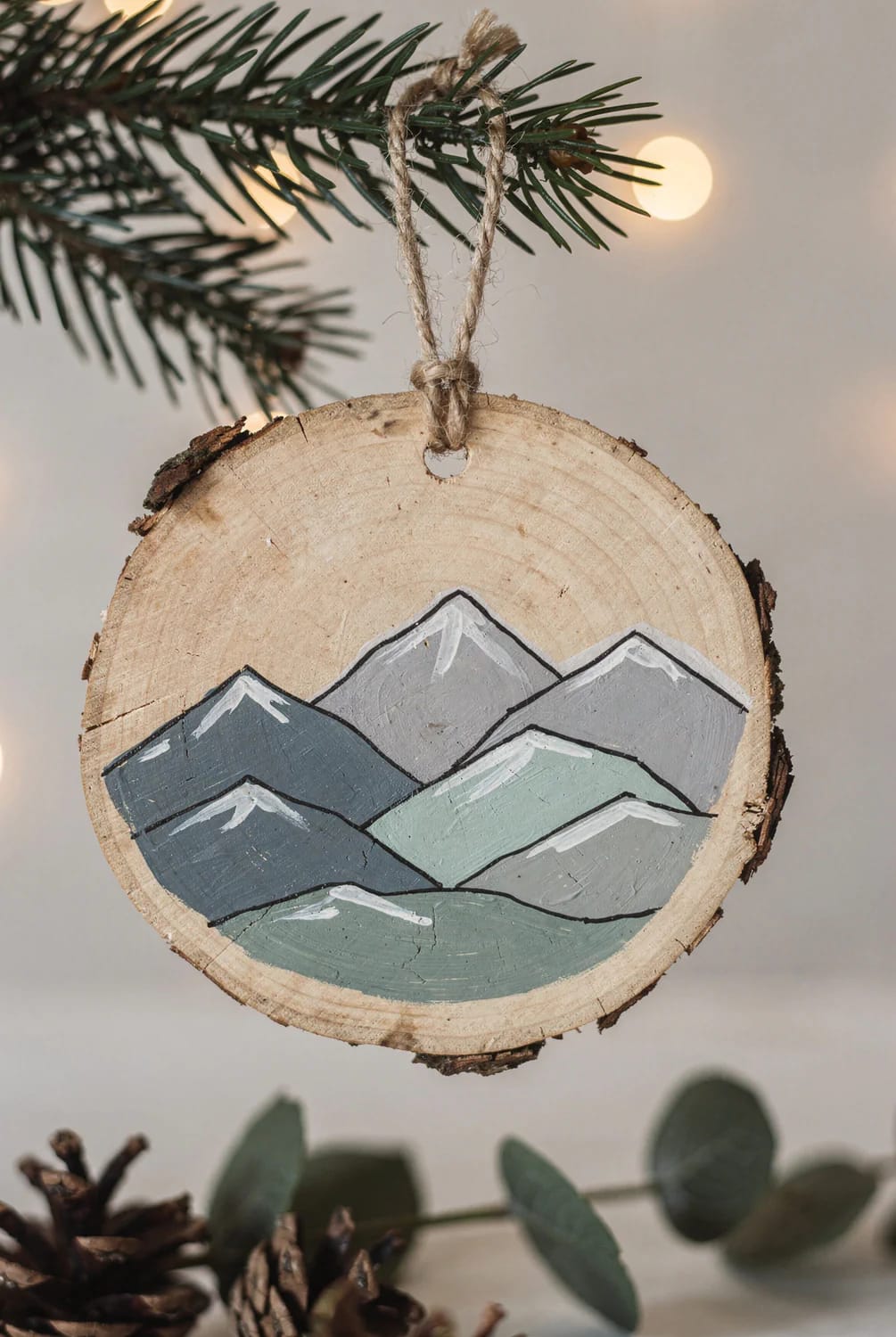 13 Painted Wood Slice Ornaments for a Minimalist Tree 6 A wood slice ornament featuring a hand-painted layered mountain range, hanging from a twine loop on a minimalist Christmas tree, with pine needles and soft lights in the background, illustrating a design from 13 painted wood slice ornaments for a minimalist tree.