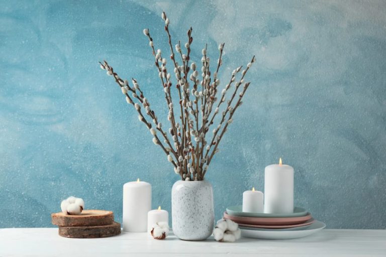 Bare Branch Winter Centerpiece Ideas for a January Table 1 A bare branch winter centerpiece featuring pussy willow branches in a speckled white vase, surrounded by white candles, stacked ceramic plates, wood slices, and cotton accents, styled on a white table against a blue textured background for a January table setting.