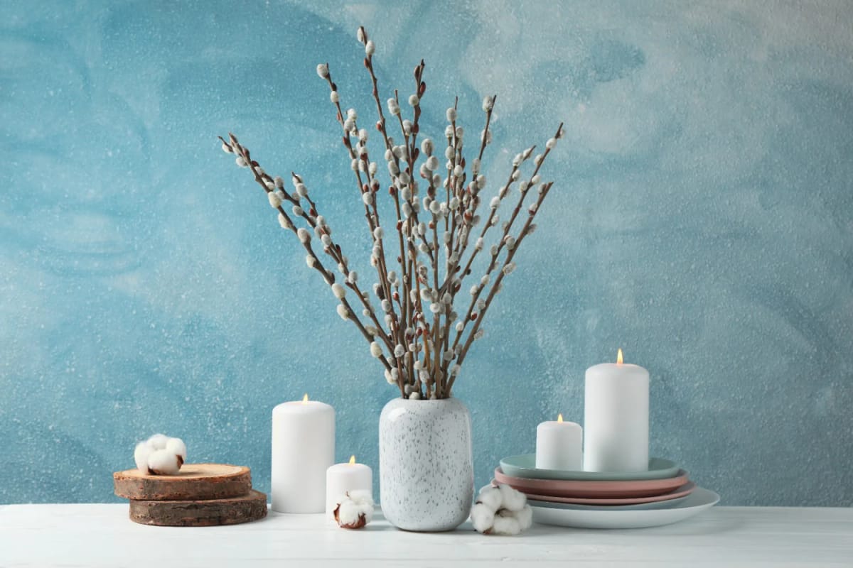 Home Decor 18 A bare branch winter centerpiece featuring pussy willow branches in a speckled white vase, surrounded by white candles, stacked ceramic plates, wood slices, and cotton accents, styled on a white table against a blue textured background for a January table setting.