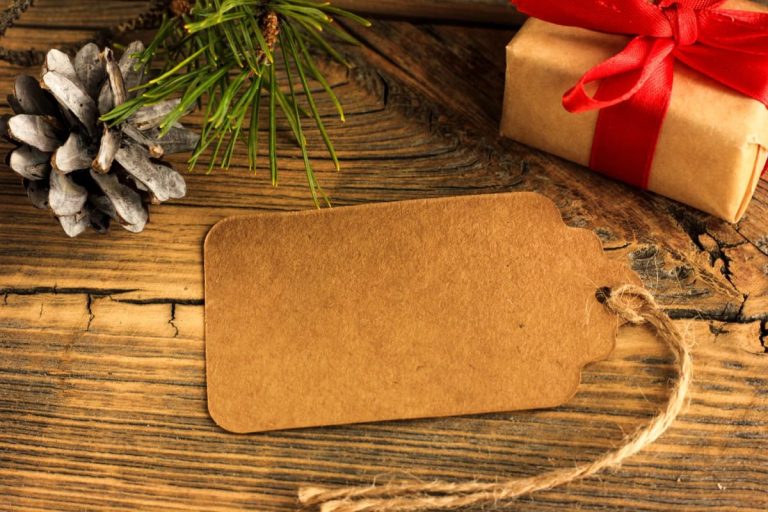 A brown paper card with twine lies on rustic wood beside a wrapped gift with a red ribbon and a pinecone, illustrating a brown paper and twine Christmas place card setup.