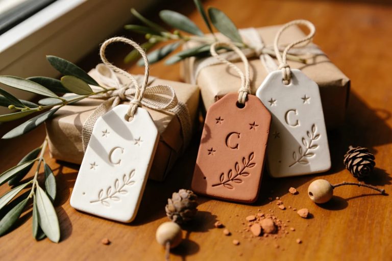 Three stamped clay gift tags in white and terracotta tones hang from wrapped kraft-paper gifts with natural twine; the tags feature stars, initials, and leafy designs, with olive branches and small pinecones on a wooden table