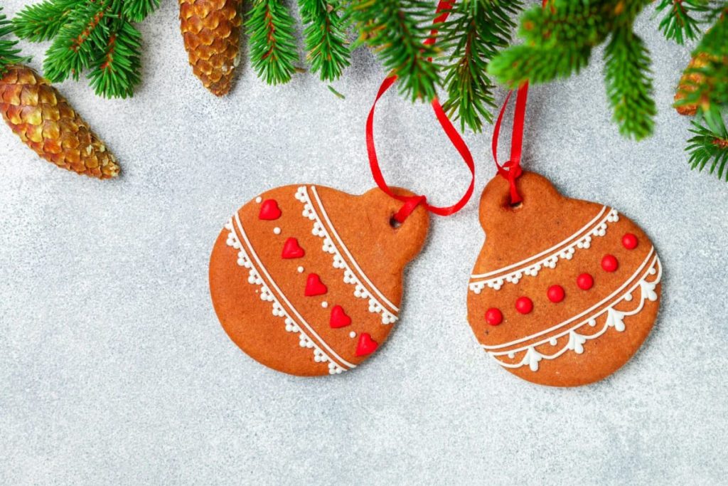 Two terracotta clay Christmas ornaments with red and white patterns hang from red ribbons, surrounded by green pine branches and pinecones, fitting an earthy tree theme.