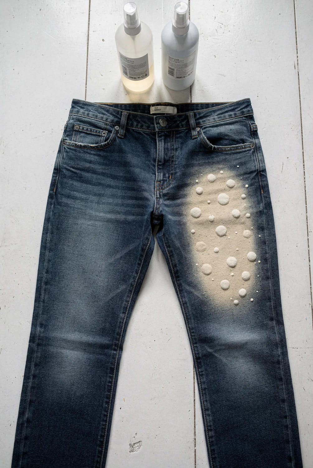 Bleach Splatter Jeans With Layered Depth Dark blue jeans lie on a light floor with a concentrated cluster of pale bleach splatters across the upper right thigh and two spray bottles positioned above them, illustrating deliberate bleach splatter jeans with varied spot sizes