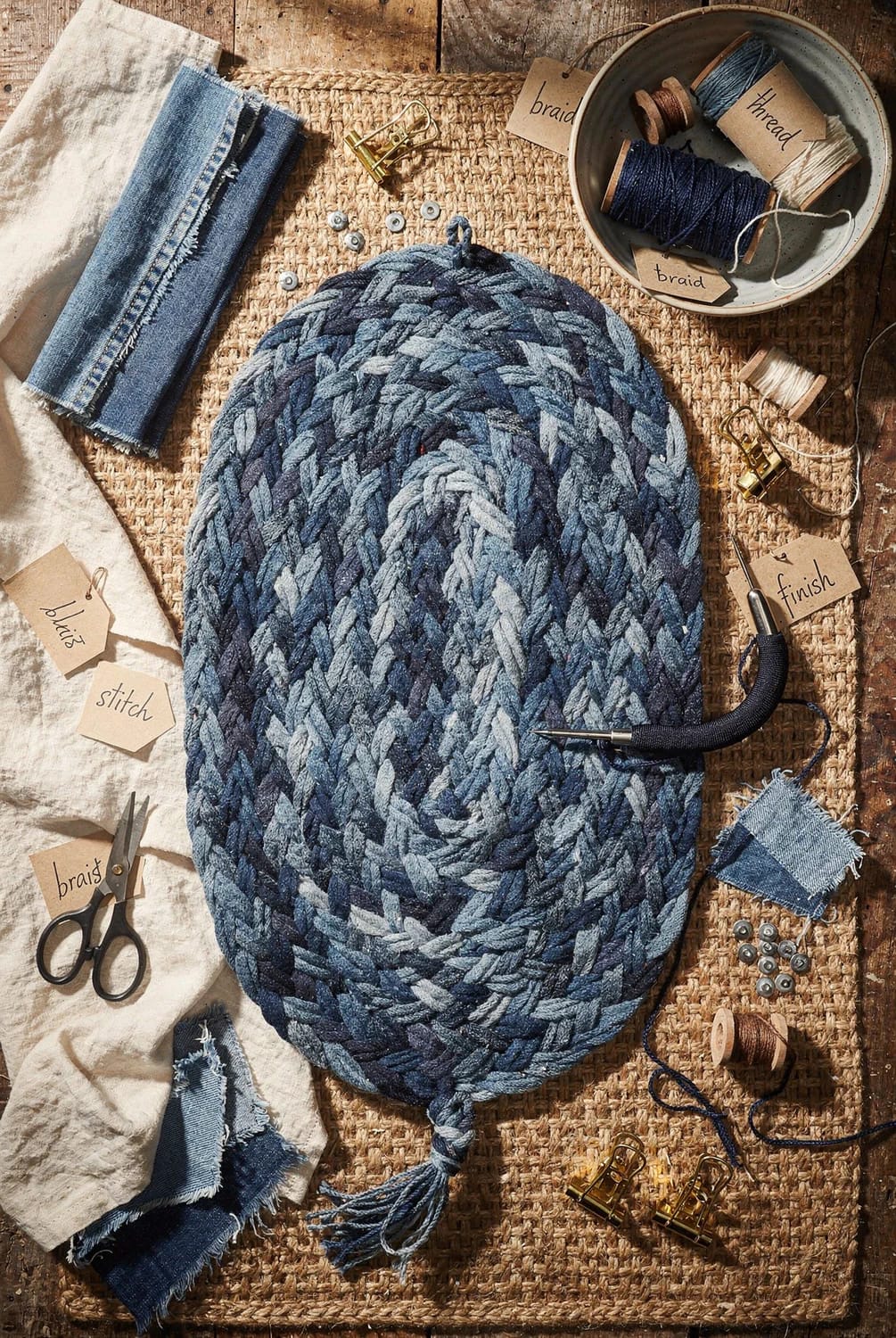 How to Make a Braided Denim Rag Rug From Old Jeans 4 Oval braided denim rag rug made from old jeans, coiled into shape on a woven work surface with thread, fabric scraps, clips, scissors, and a needle nearby
