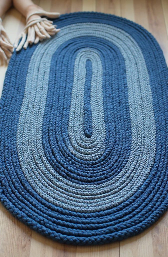 How to Make a Braided Denim Rag Rug From Old Jeans 5 Oval braided denim rag rug in alternating blue shades, coiled into concentric rows on a wooden floor with a hand holding braided fabric at the upper left.