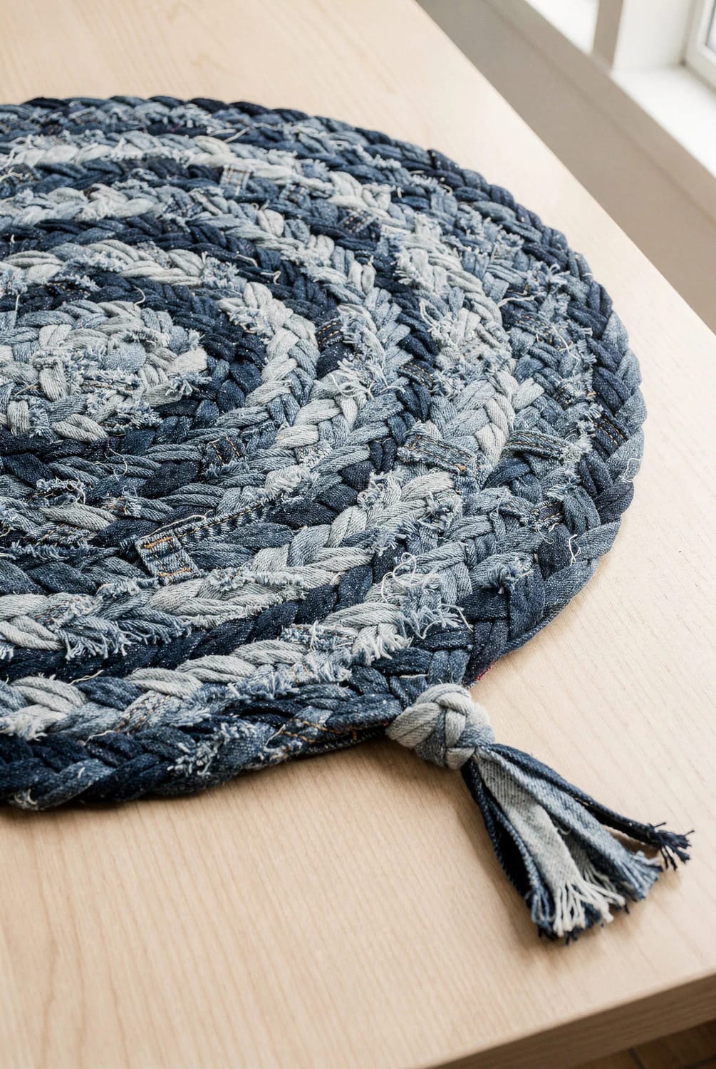 Close-up of a braided denim rag rug made from old jeans, with its narrow braided tail extending onto a light wood surface