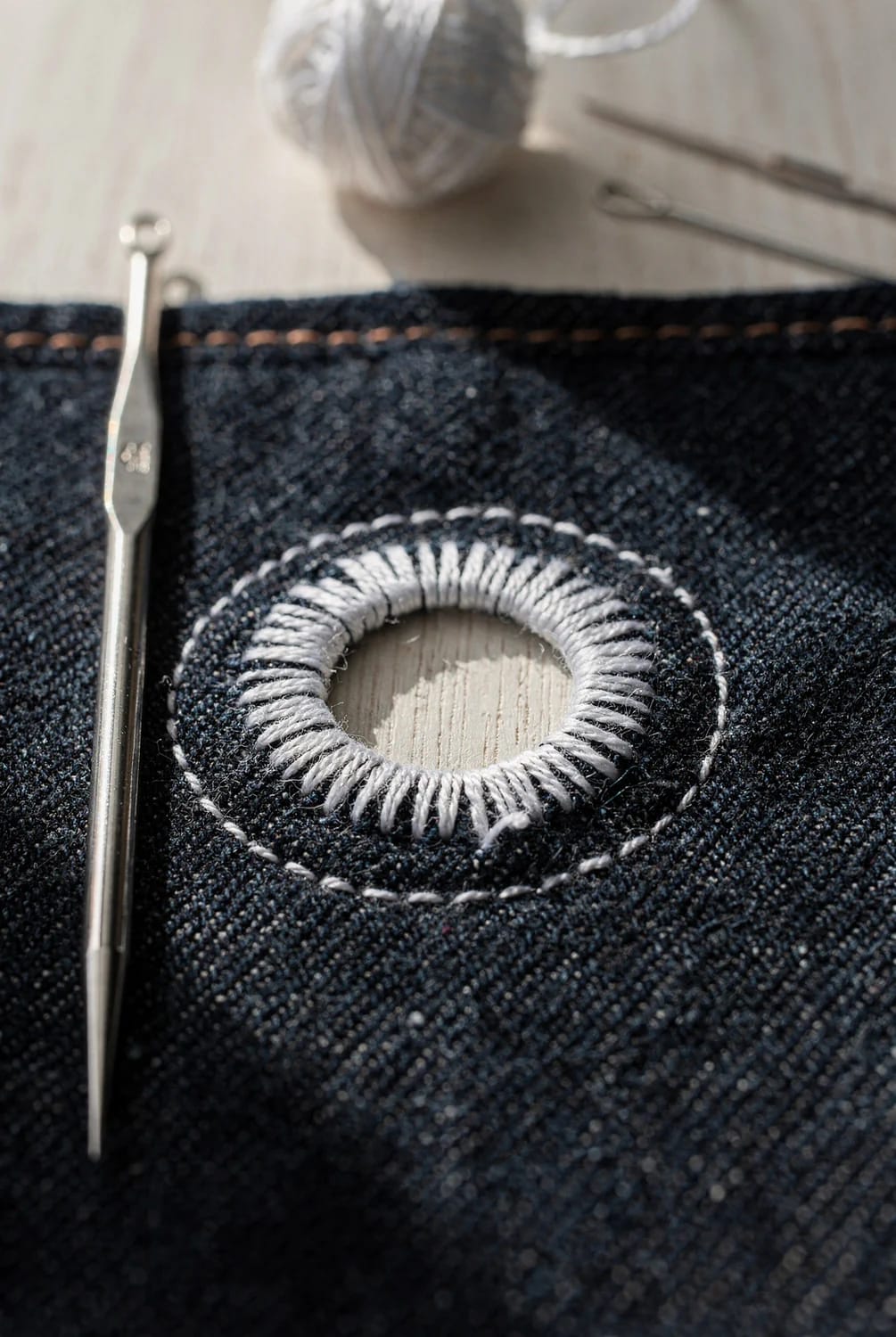 Broderie Anglaise Jeans Eyelet Stitching Close-up of a white embroidered eyelet ring on dark denim, with a metal needle beside it and thread visible in the background, illustrating Broderie Anglaise jeans technique