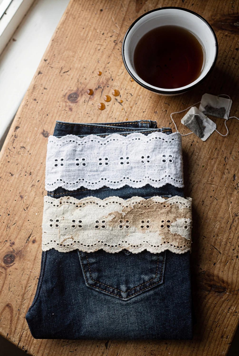 Broderie Anglaise Jeans With Eyelet Lace Dark blue jeans folded on a wooden workbench with white and cream broderie anglaise lace bands across the back pocket, beside a cup of tea and used tea bags