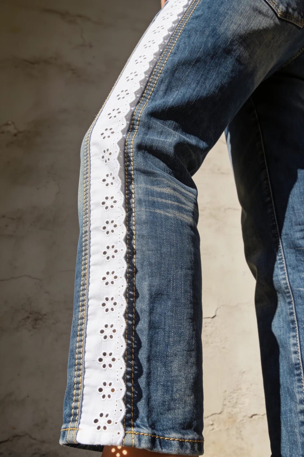 Broderie Anglaise Jeans With Seam-Set Eyelet Panel Close-up of blue jeans featuring a white broderie anglaise eyelet panel sewn into the outer leg seam, with yellow topstitching and a scalloped edge