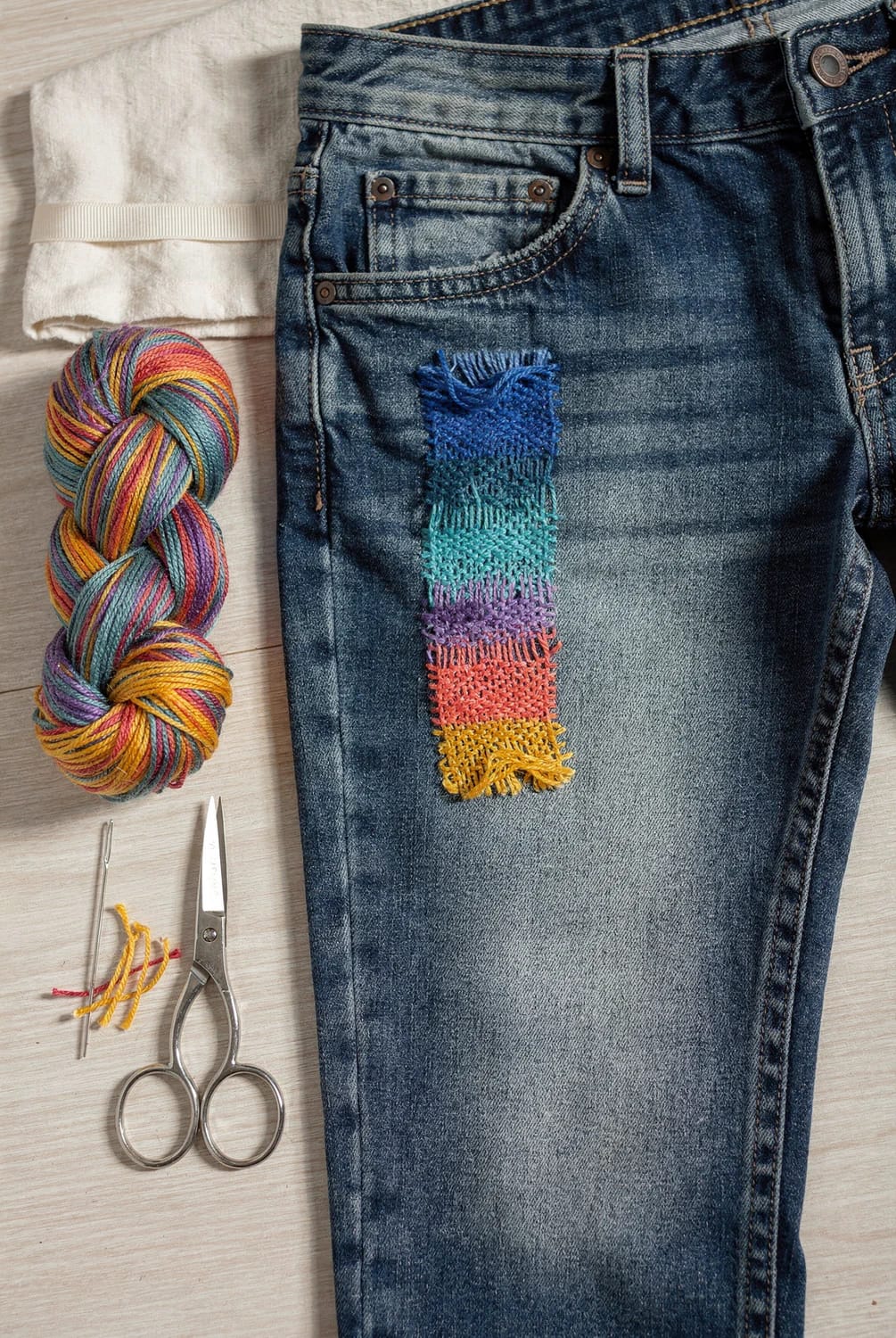 Colorful Thread Darning Gradient On Jeans Blue jeans with a rectangular gradient darn in bright blue, teal, purple, coral, and yellow stitches, beside a variegated multicolor skein, needle, scissors, and loose threads for colorful thread darning.
