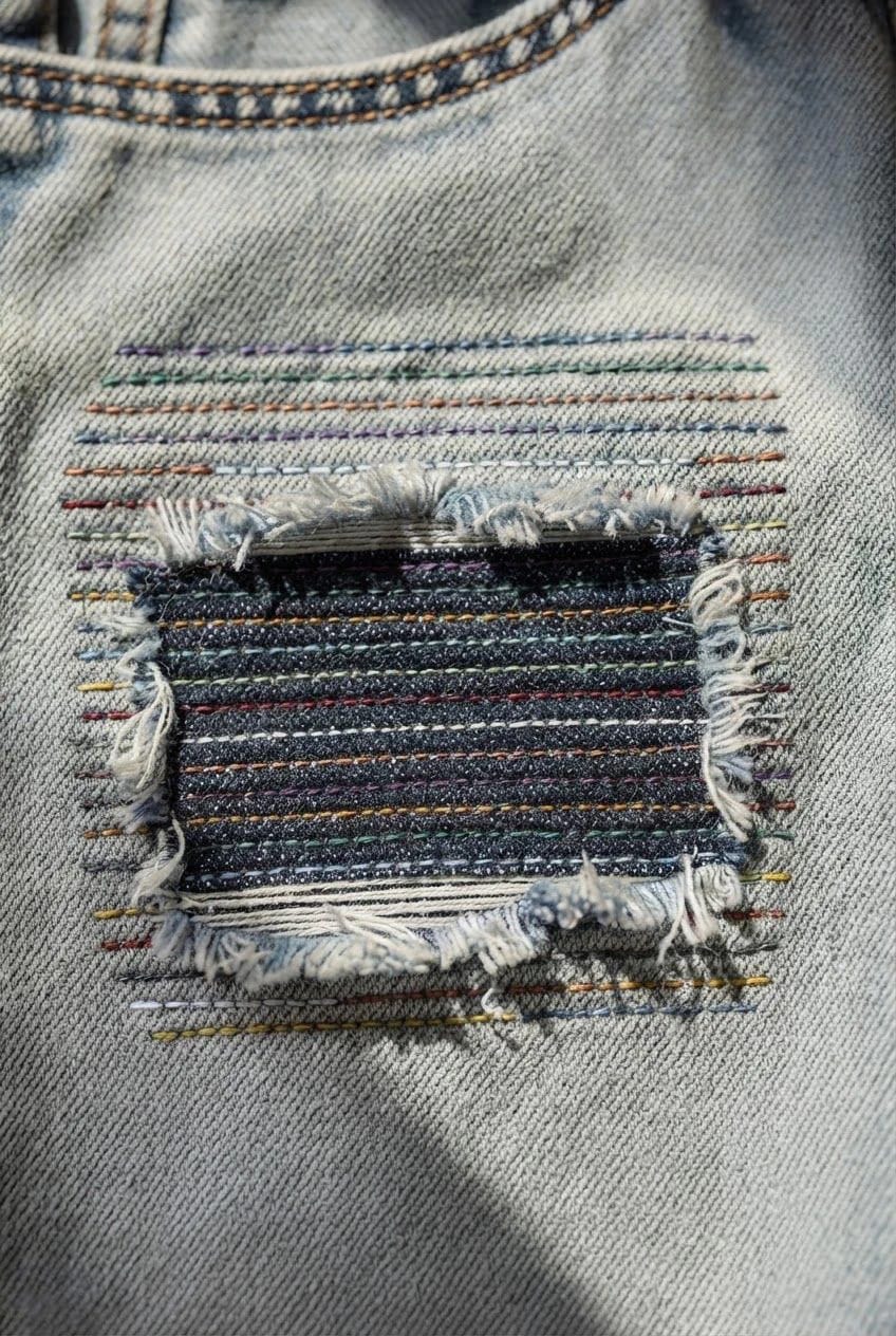 Colorful Thread Darning On Jeans Close-up of jeans repaired with a dark stacked boro patch, frayed edges, and rows of colorful horizontal stitches across the denim
