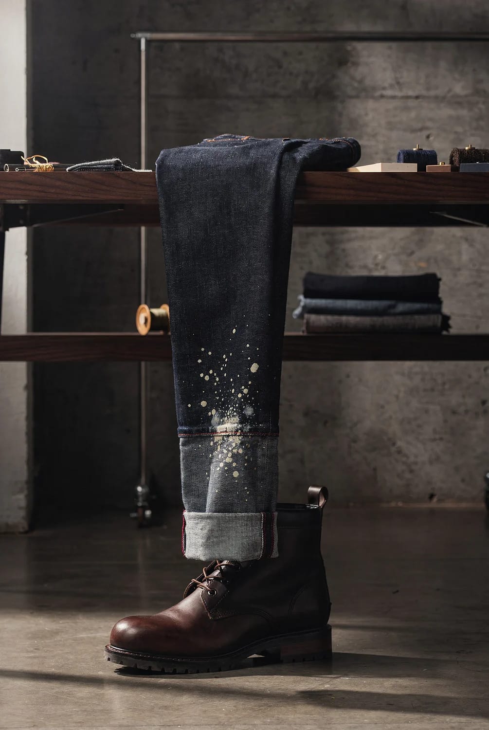 Deliberate Bleach Splatter Jeans With Turn-Up Dark jeans draped over a wooden workbench, featuring concentrated light bleach splatter around the lower leg and folded turn-up above a brown-and-black work boot, illustrating deliberate bleach splatter jeans styling.