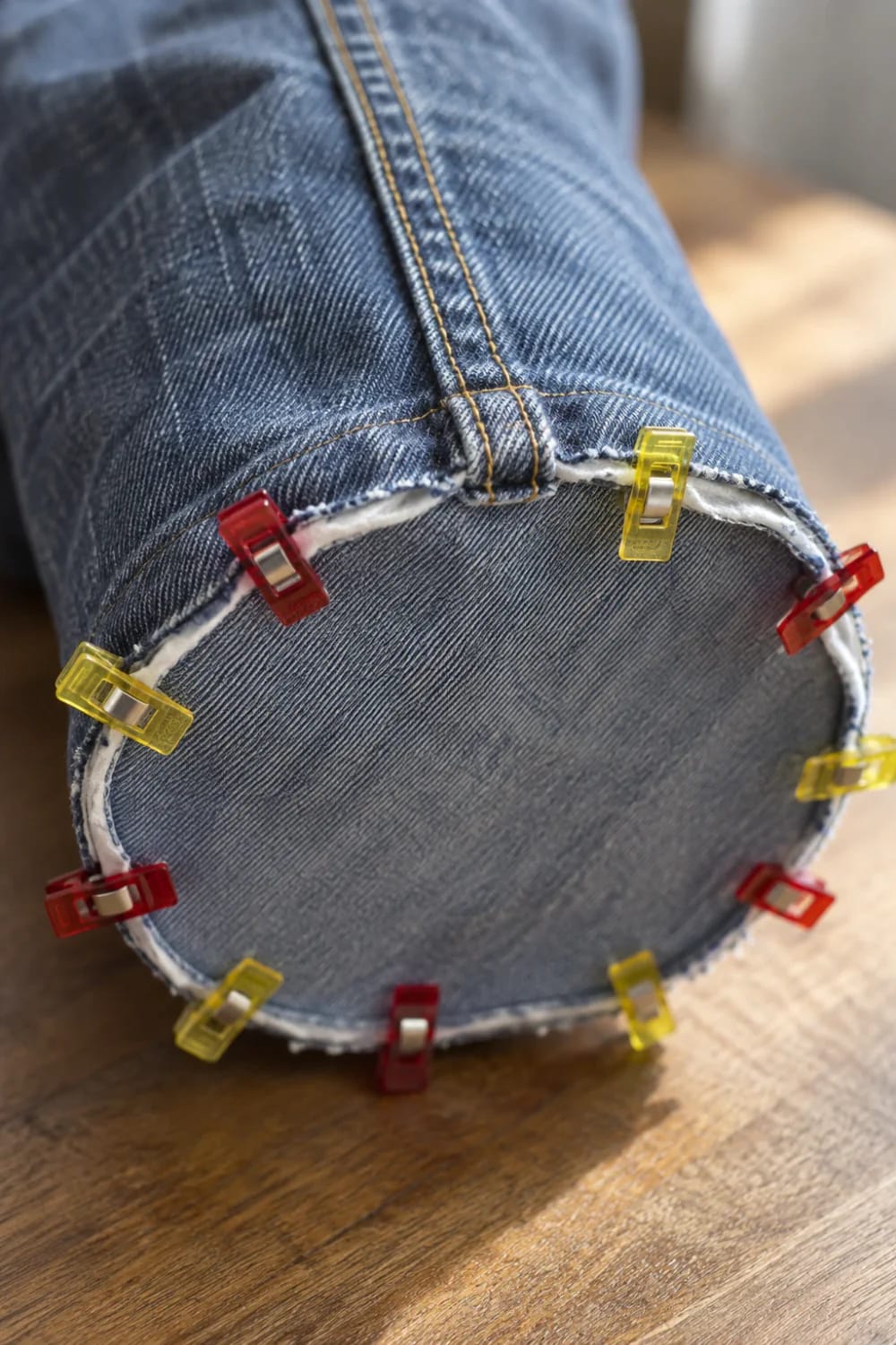 Denim Bucket Bag Base Sewing Tutorial Close-up of a denim tube with a circular fabric base pinned around its edge using red and yellow sewing clips, illustrating how to sew the base into a denim bucket bag