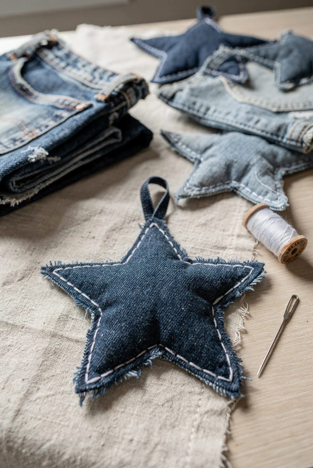 Denim Fabric Star Ornaments With Sashiko Stitching A dark denim star ornament outlined with white sashiko stitching lies on fabric, surrounded by additional denim stars, old jeans, white thread, and a needle.