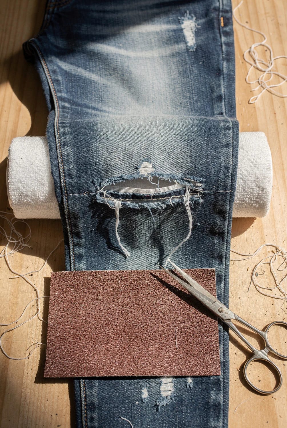 Distressed Jeans With Frayed Knee Slit Distressed blue jeans with a large frayed knee slit rest on a rolled towel, alongside sandpaper, scissors, and loose white threads on a wooden surface