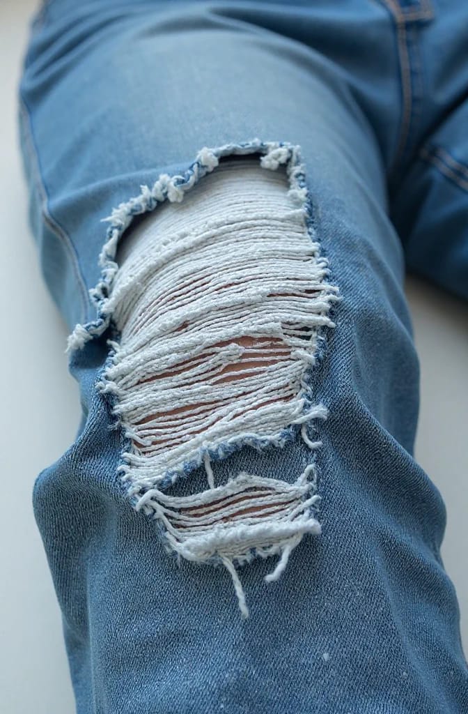 Distressed Jeans With Frayed Knee Slits Close-up of blue distressed jeans showing large frayed knee slits cut across the leg, with white horizontal threads forming a ladder-like texture