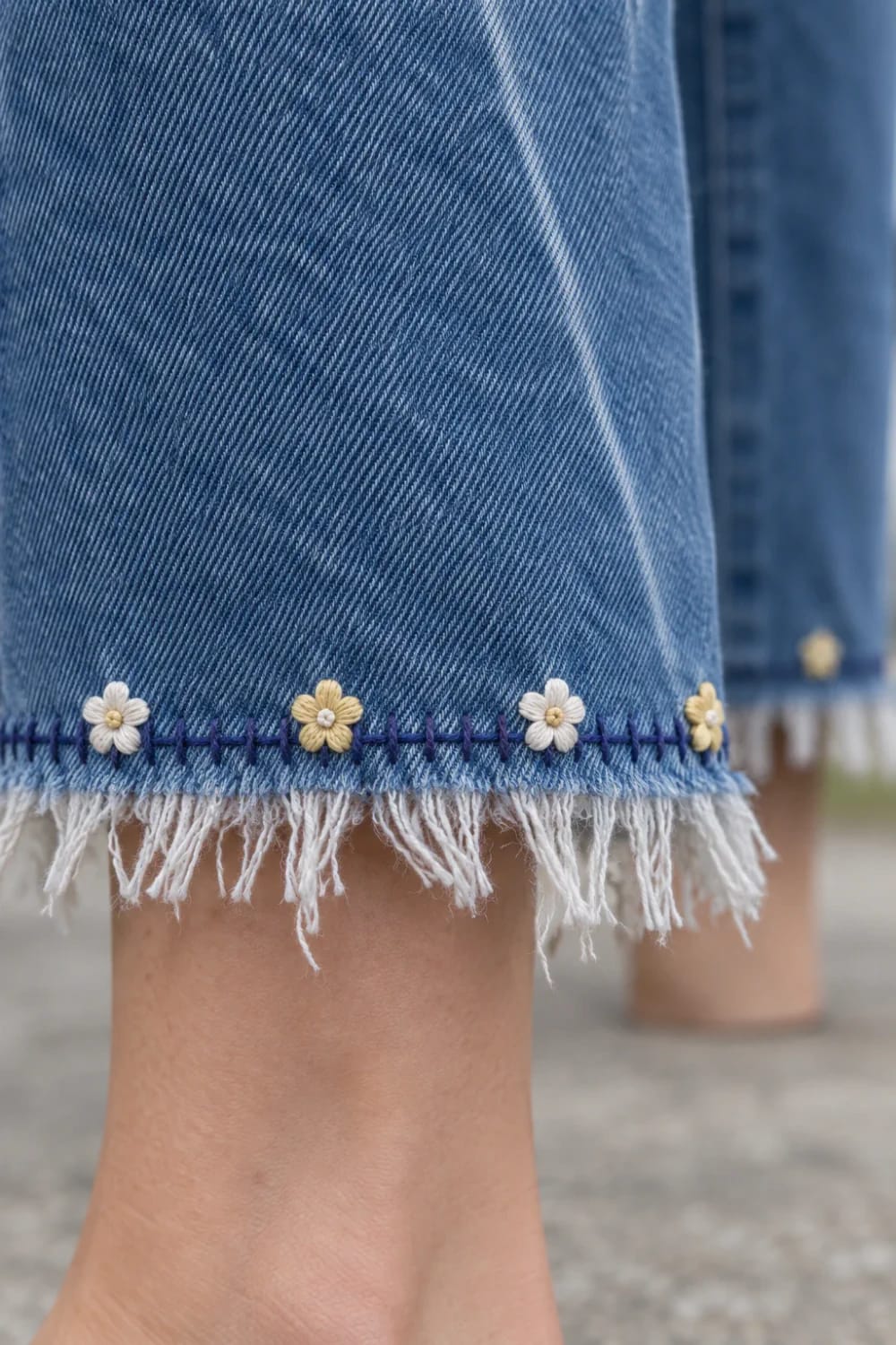 Floral Embroidered Hem Jeans Ideas Close-up of blue jeans with a frayed hem decorated with small white and yellow embroidered flowers and purple stitching, illustrating floral embroidered hem jeans ideas