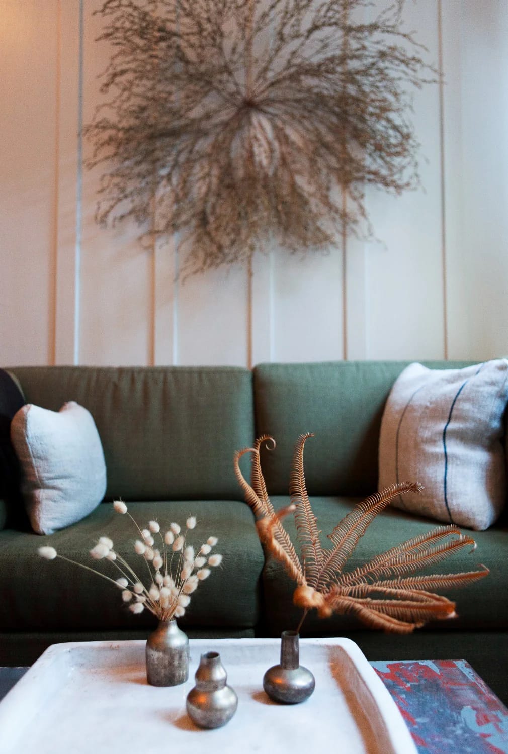 Three small metallic vases holding dried decorative stems sit on a white living room centre table, with a green sofa and cushions in the background, illustrating a best out of waste showpiece idea