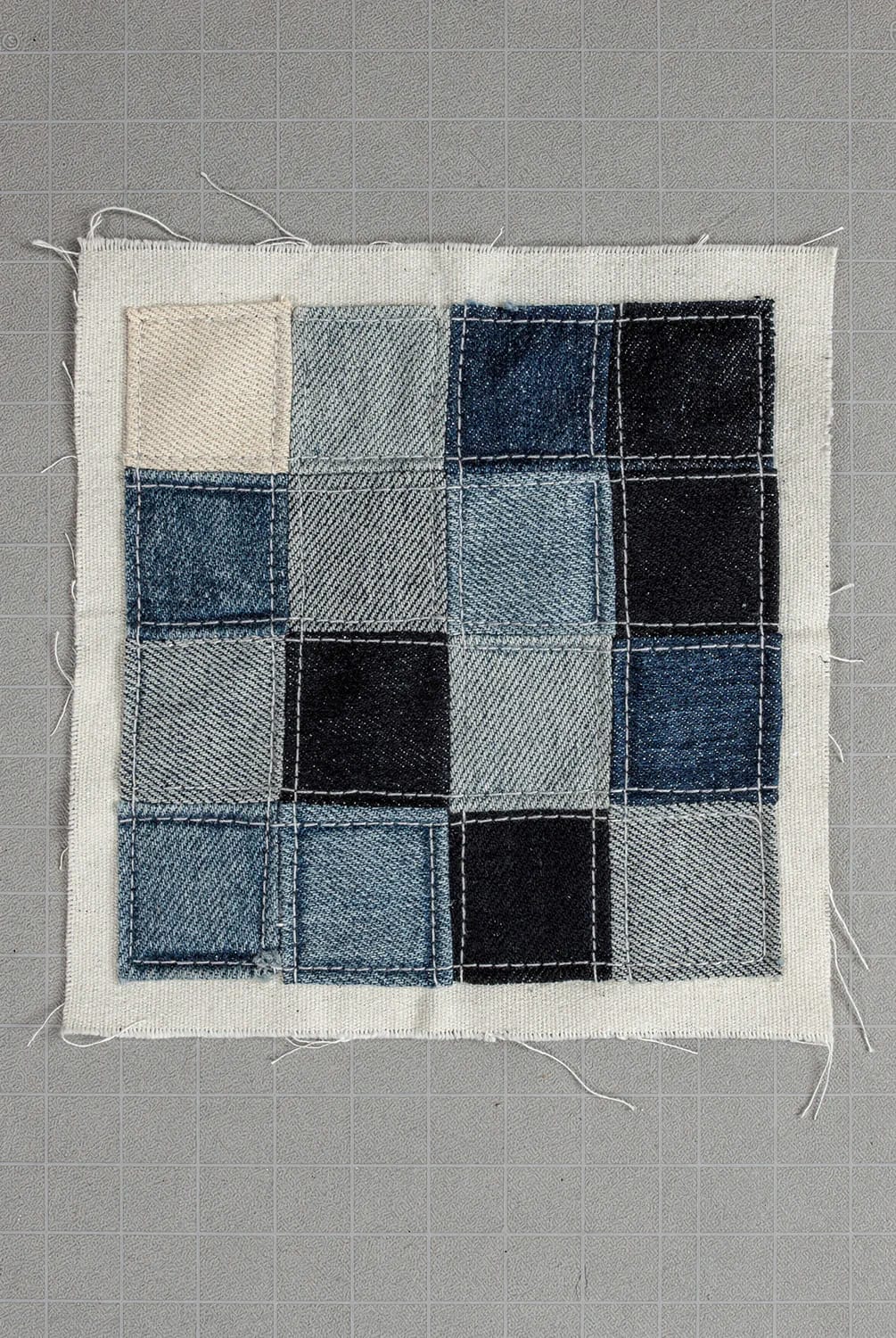 Boro Patched Jeans Scrap Layering Sample A rectangular fabric panel displays sixteen stitched denim scraps in varied blue, gray, black, and beige tones, arranged in a grid on a cutting mat for boro patched jeans repair.