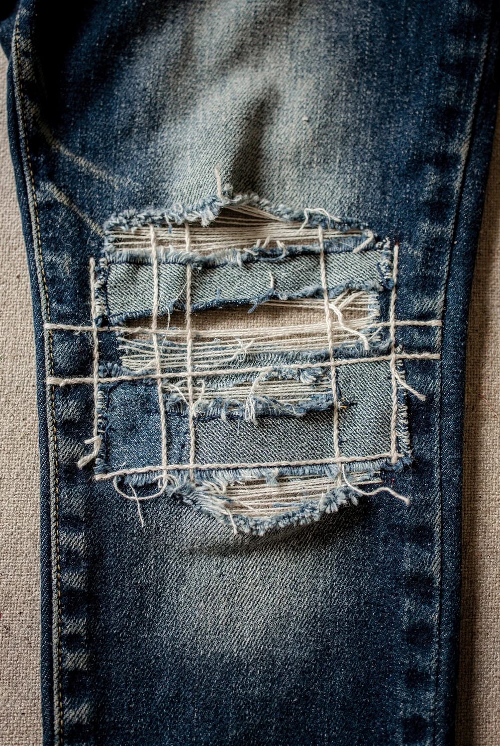 Boro Patched Jeans With Layered Denim Scraps Close-up of blue jeans repaired with overlapping denim scraps, exposed threads, and visible cross-stitching around a worn tear, illustrating layered boro patched jeans