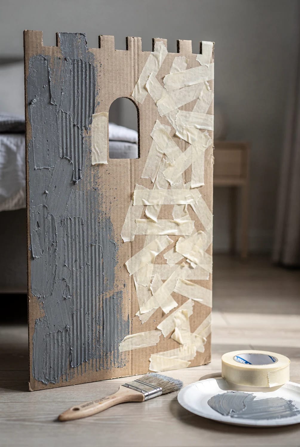 Cardboard castle playhouse panel with crenellated top and arched window, partly coated in textured gray paint and partly covered with overlapping strips of masking tape to create a stone-like finish; a paintbrush, tape roll, and plate of gray paint sit nearby