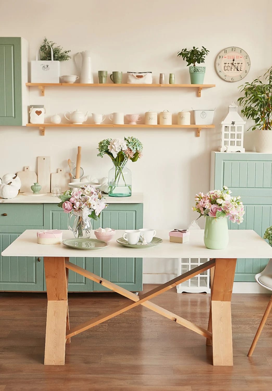 Cottage kitchen with muted green cabinets, open wooden shelves holding cups and pitchers, floral arrangements, potted plants, and a farmhouse-style table with tea settings.