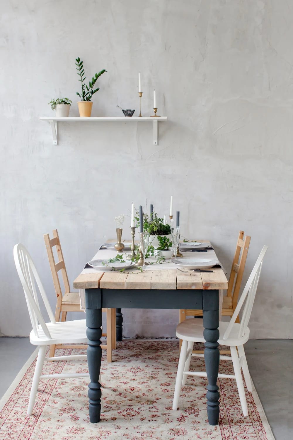 Long French farm table with a rustic wood top and dark turned legs, surrounded by mismatched white and natural wood chairs. Plates, greenery, candlesticks, and glassware create a collected junk chic cottage decor dining setting.
