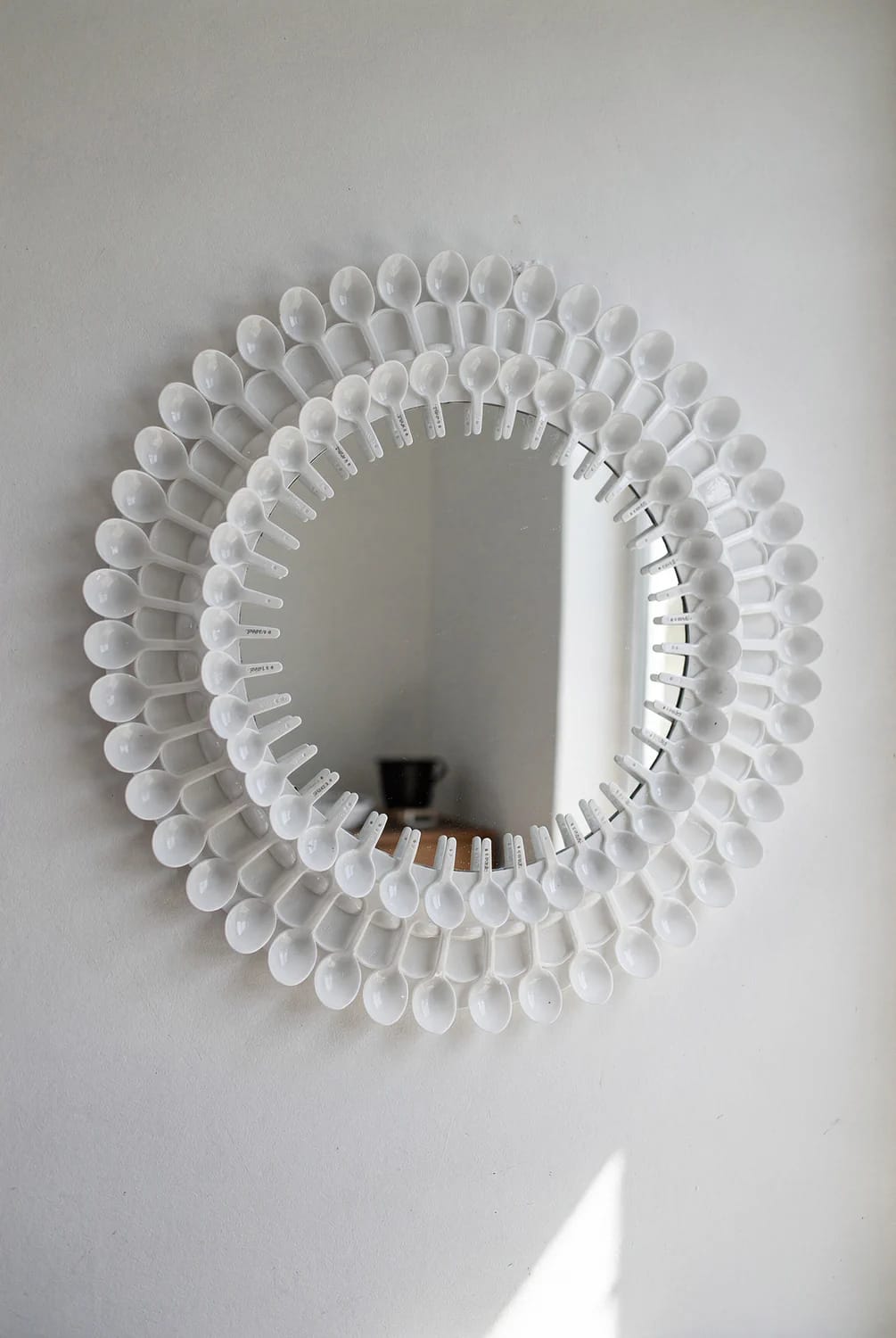 A wall mirror surrounded by concentric rows of white plastic spoons, creating a decorative junk art frame