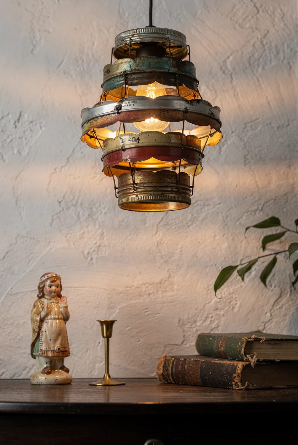 A glowing hanging lamp made from stacked, weathered metal scrap pieces appears above a table with a figurine, candlestick, and old books, illustrating a junk art lamp idea.