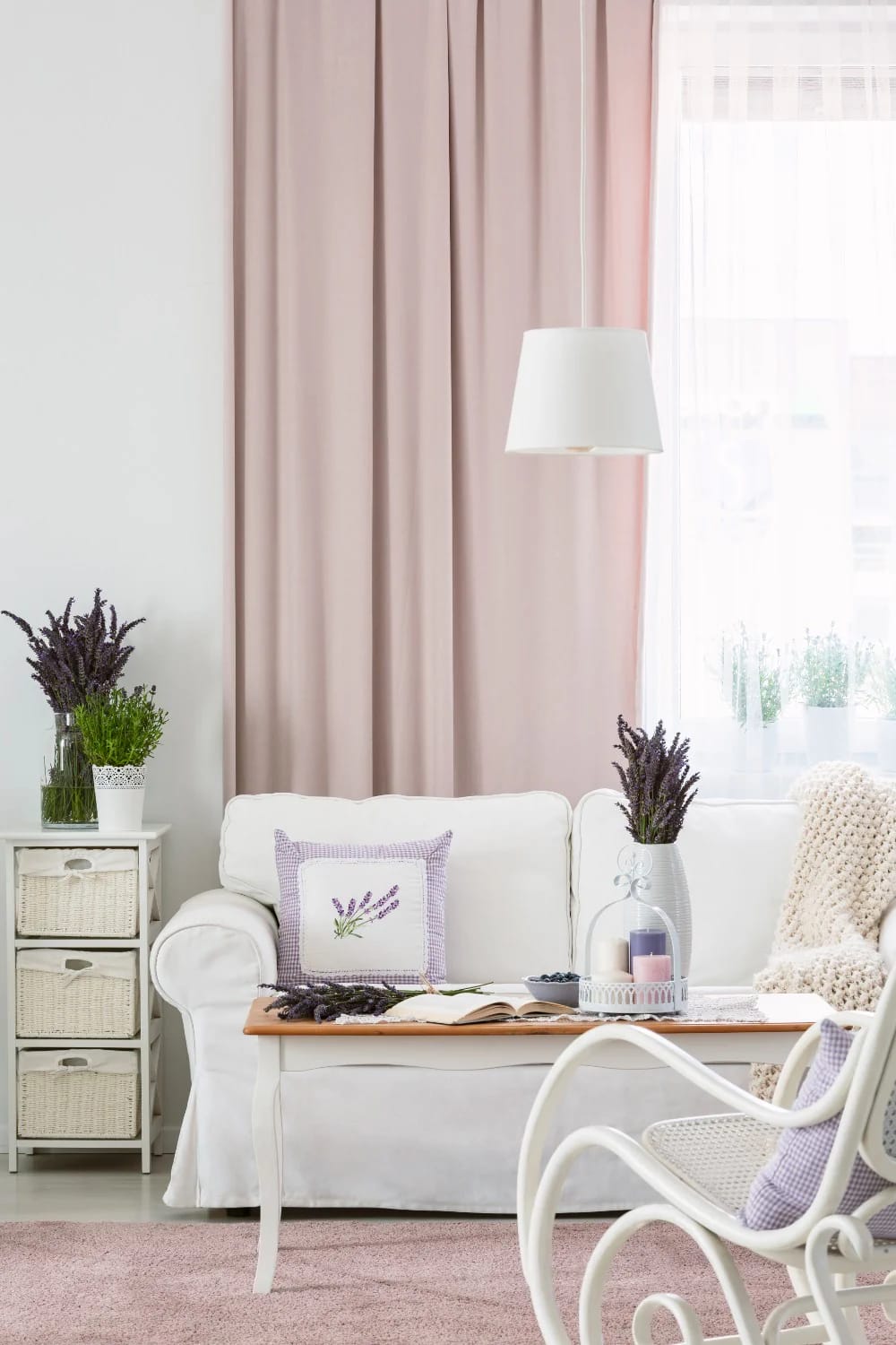 Bright cottage living room with creamy white walls, a white sofa, blush-pink curtains and rug, lavender floral arrangements, a small coffee table, and white wicker seating.