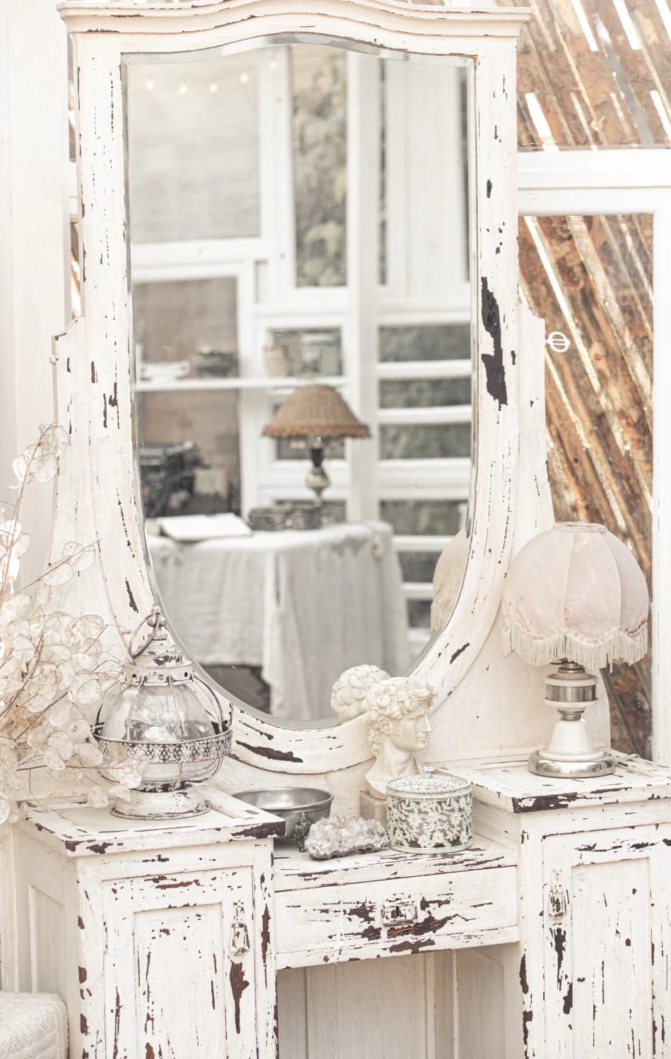 Junk chic cottage decor featuring a distressed white vanity with a large mirror, chipped paint, a small lamp, decorative containers, and a glass lantern.