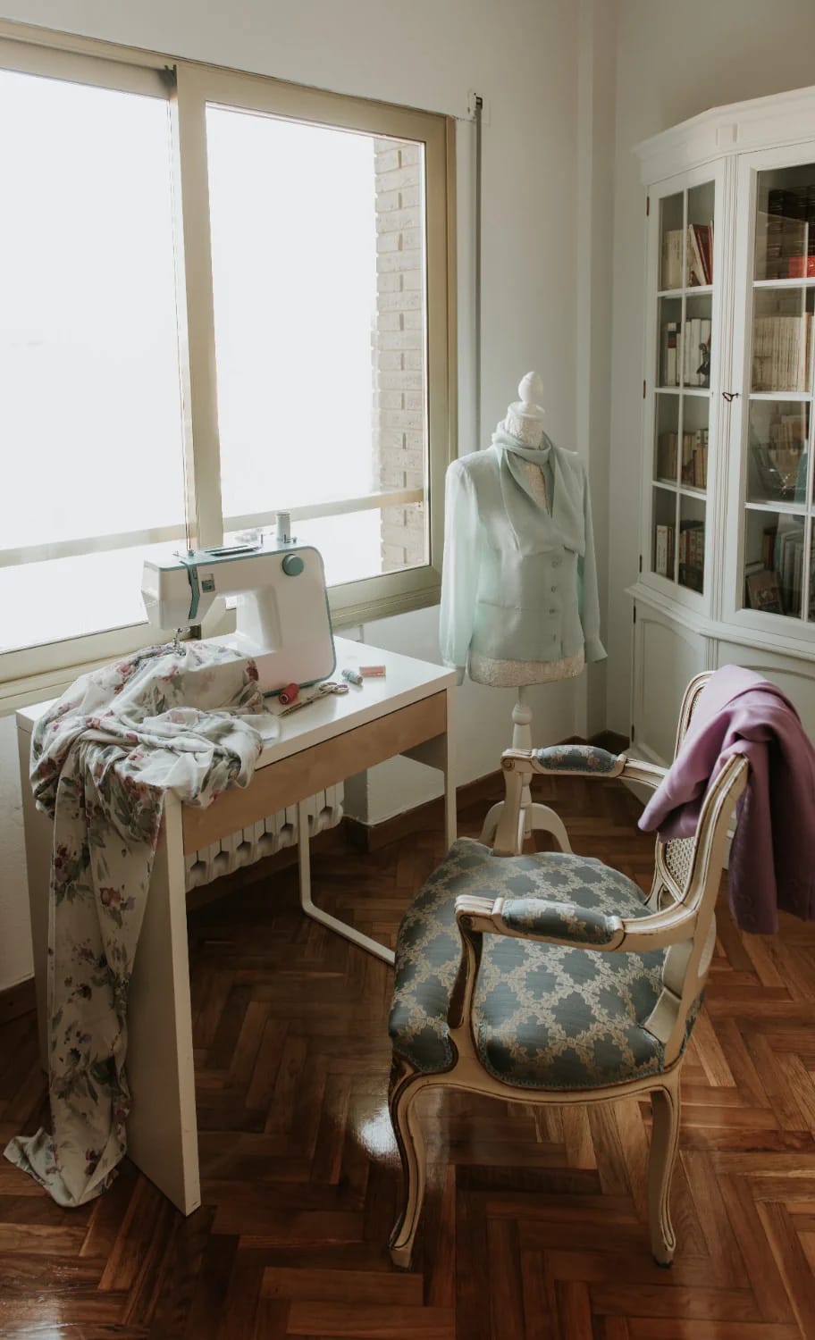Junk chic cottage sewing corner with a sewing machine, floral fabric, dress form, upholstered vintage chair, and glass-front bookcase beside a bright window