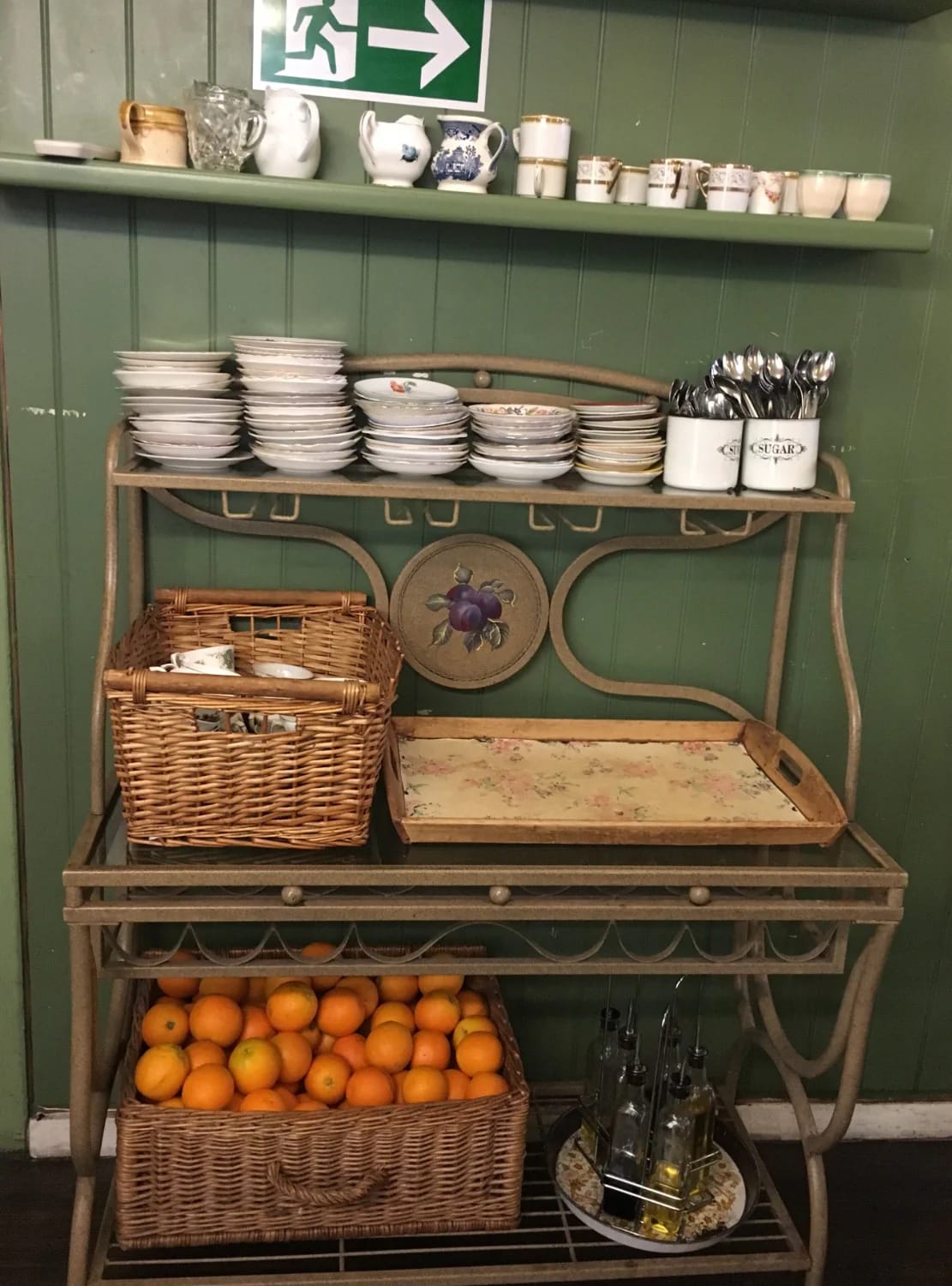 Junk chic cottage decor features a vintage metal sideboard holding stacked dishes, cups, pitchers, wicker baskets, oranges, bottles, and serving utensils against a dark green paneled wall