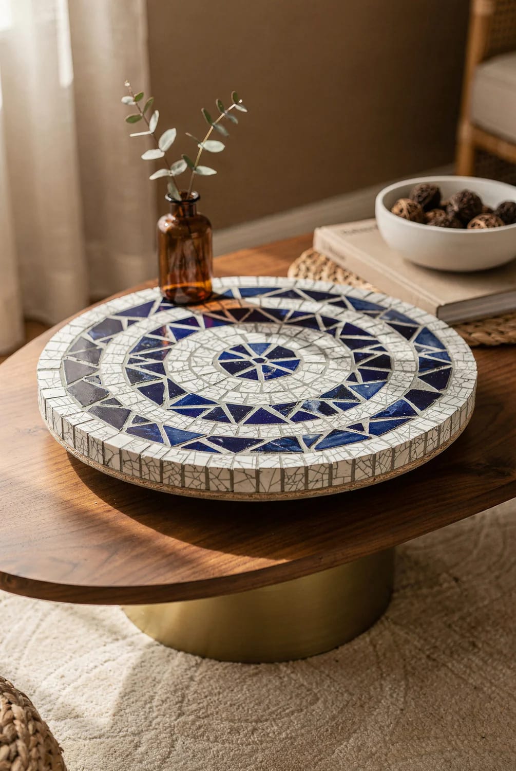 Round blue and white mosaic showpiece displayed on a wooden living room centre table, beside an amber vase with leafy stems and a bowl of decorative balls