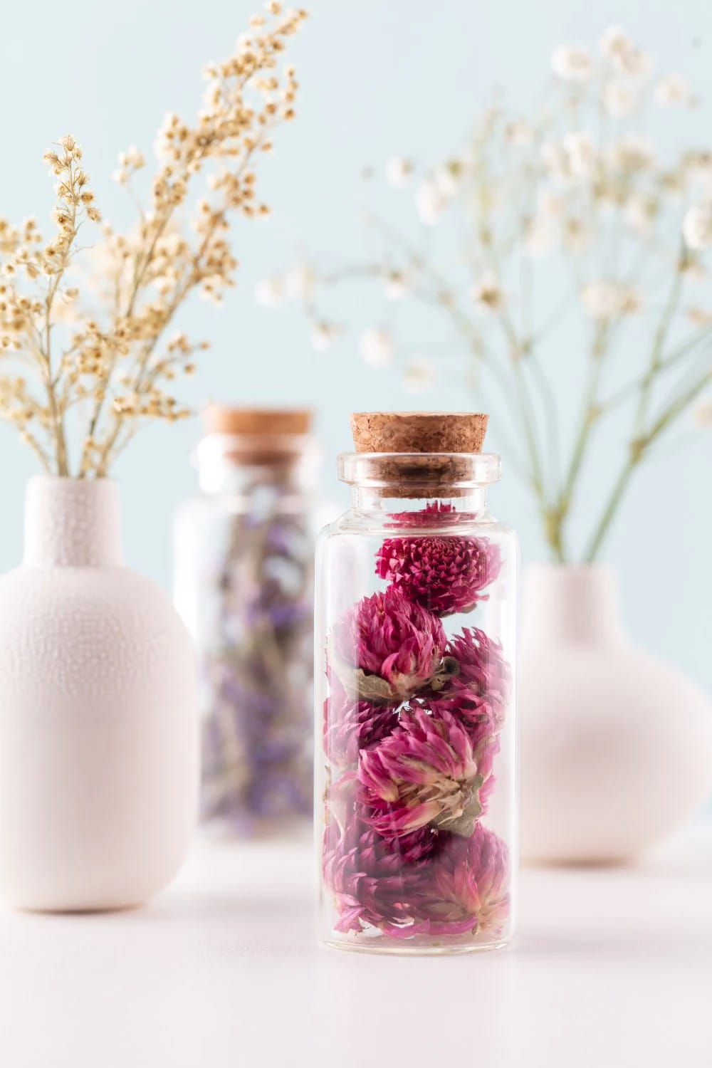 Clear glass jar with a cork lid containing dried pink flowers, displayed among pale decorative vases as an out of waste showpiece for a living room centre table