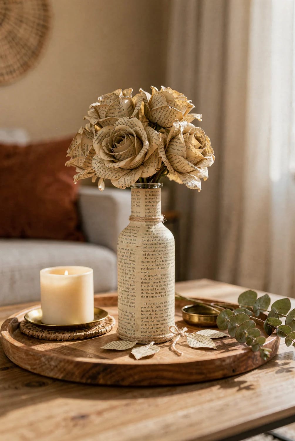 Paper roses made from printed pages arranged in a tall matching paper-covered vase on a wooden centre table, beside a lit candle and greenery.
