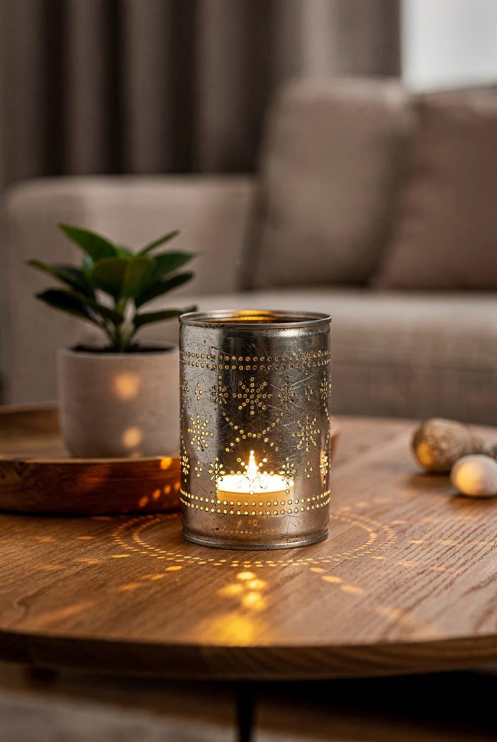 Perforated metal candle holder glowing on a wooden living room centre table, casting patterned light beside a potted plant.