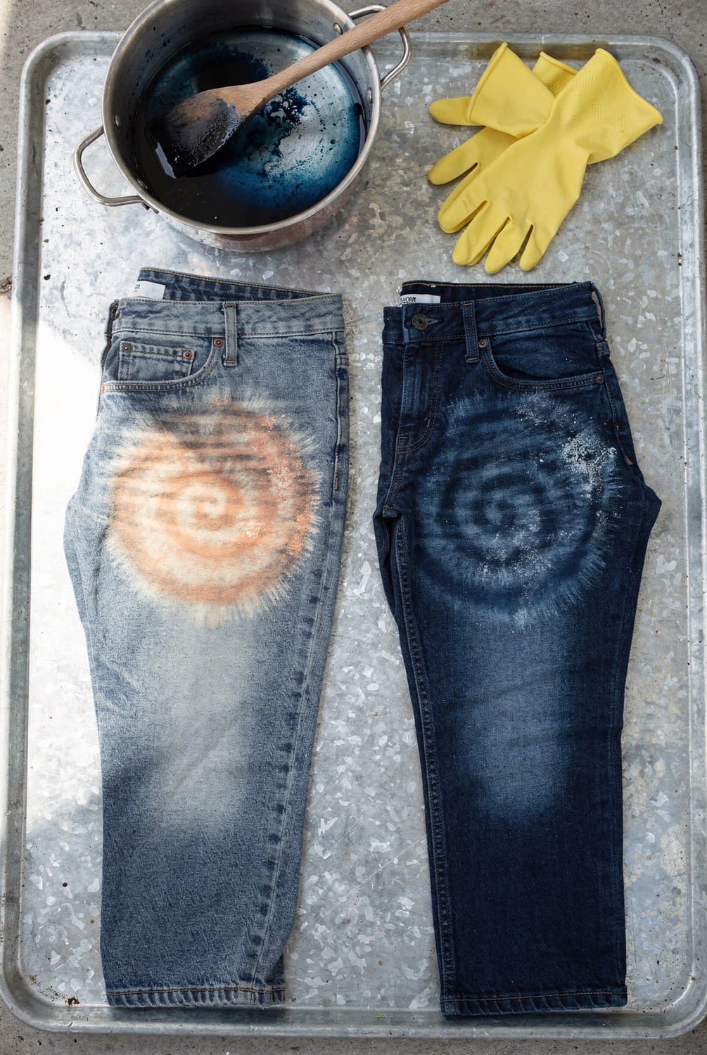 Reverse Tie Dye Jeans With Bleach Two pairs of jeans with circular bleach patterns rest on a metal tray beside a pot of dark dye, a wooden spoon, and yellow rubber gloves for reverse tie dye jeans with bleach