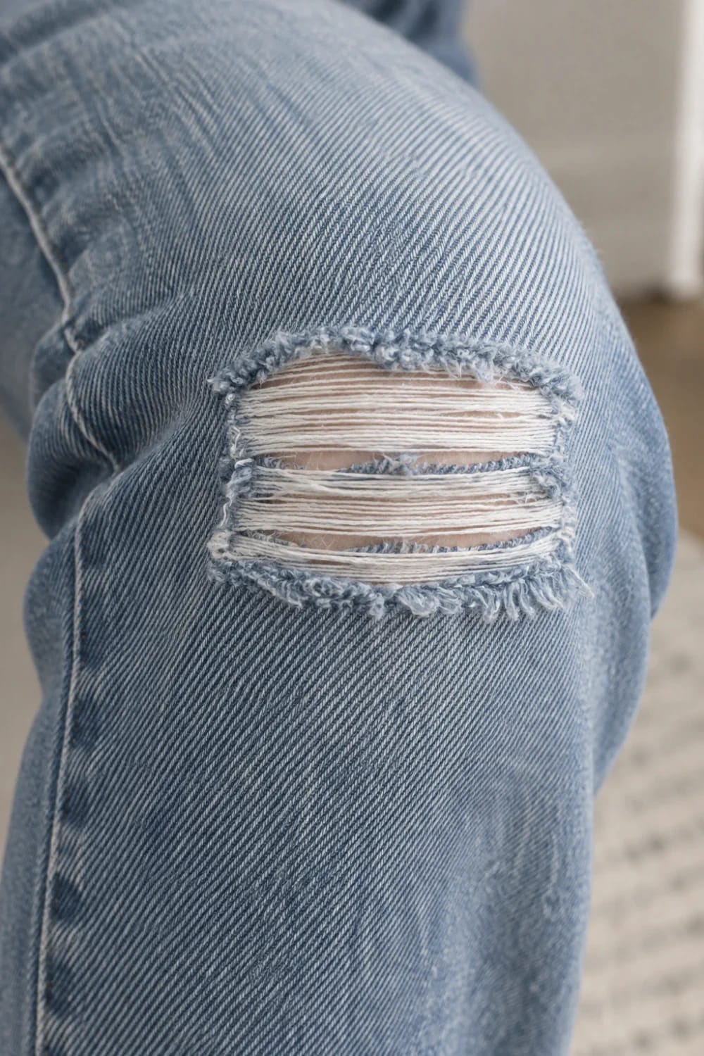 Close-up of light blue denim jeans with a rectangular shredded knee opening, showing horizontal white weft threads between frayed blue edges for a ladder effect