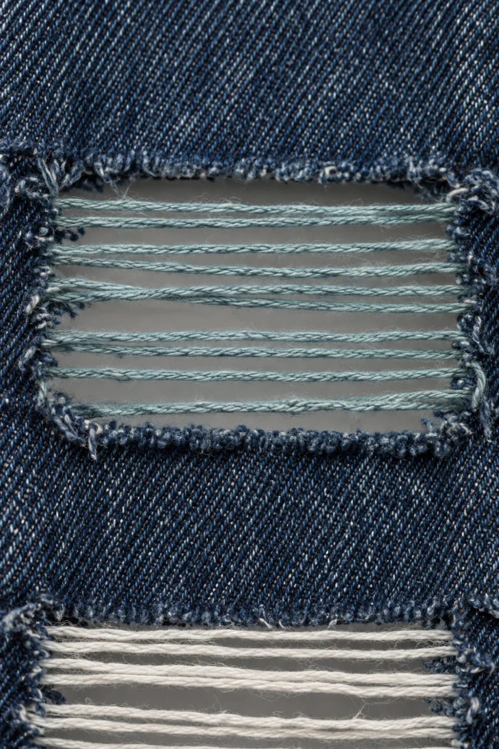 Close-up of dark blue denim with two horizontal ladder-style distressed openings; the upper opening has pale blue-gray wefts and the lower opening has bright white wefts, illustrating how to tint white wefts in shredded ladder knee jeans